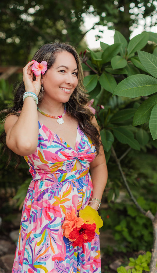 Woman wearing the colorful Aloha Rainbow Necklace with genuine gemstones and a carved mother-of-pearl hibiscus pendant in a sunlit tropical garden.
