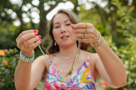 Charlotte’s Cove founder Ashley examining layered beaded and gold chain necklaces outdoors while wearing colorful bracelet stacks.