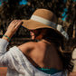 Woman wearing a wide-brimmed hat with a white band, standing outdoors with palm trees in the background. wearing the Afterglow Abalone Bracelet with abalone coin, amazonite beads, in gold plated pvd 