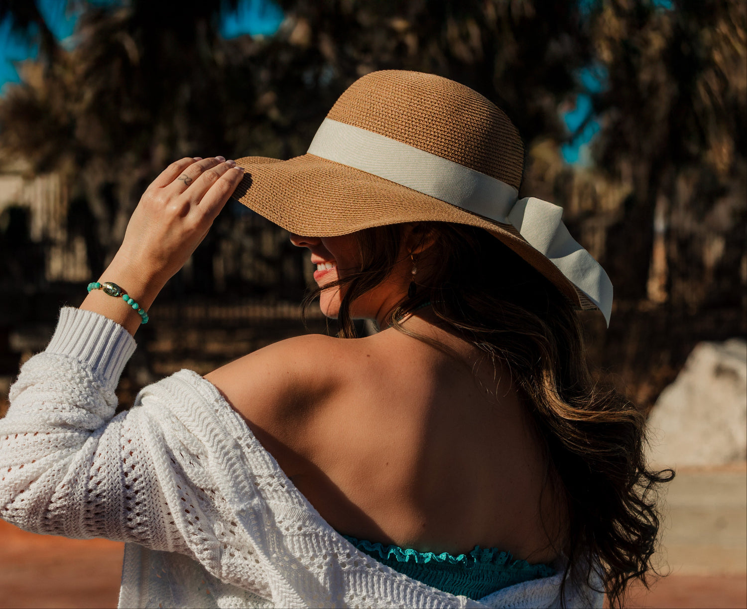 Woman wearing a wide-brimmed hat with a white band, standing outdoors with palm trees in the background. wearing the Afterglow Abalone Bracelet with abalone coin, amazonite beads, in gold plated pvd 