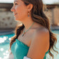 A woman in profile wearing a turquoise strapless top and white knit cardigan, with matching turquoise jewelry, photographed outdoors near a pool.
