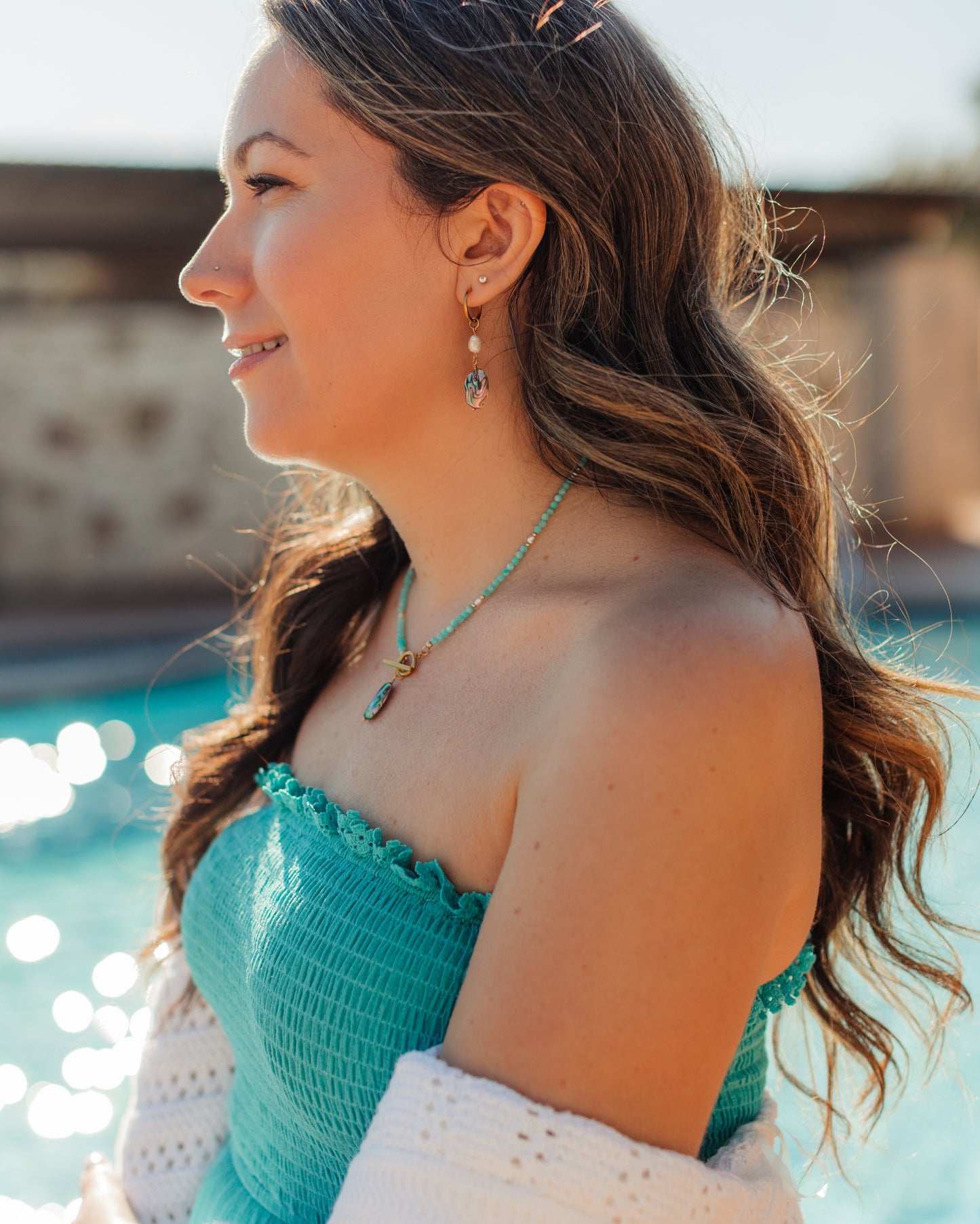 A woman in profile wearing a turquoise strapless top and white knit cardigan, with matching turquoise jewelry, photographed outdoors near a pool.
