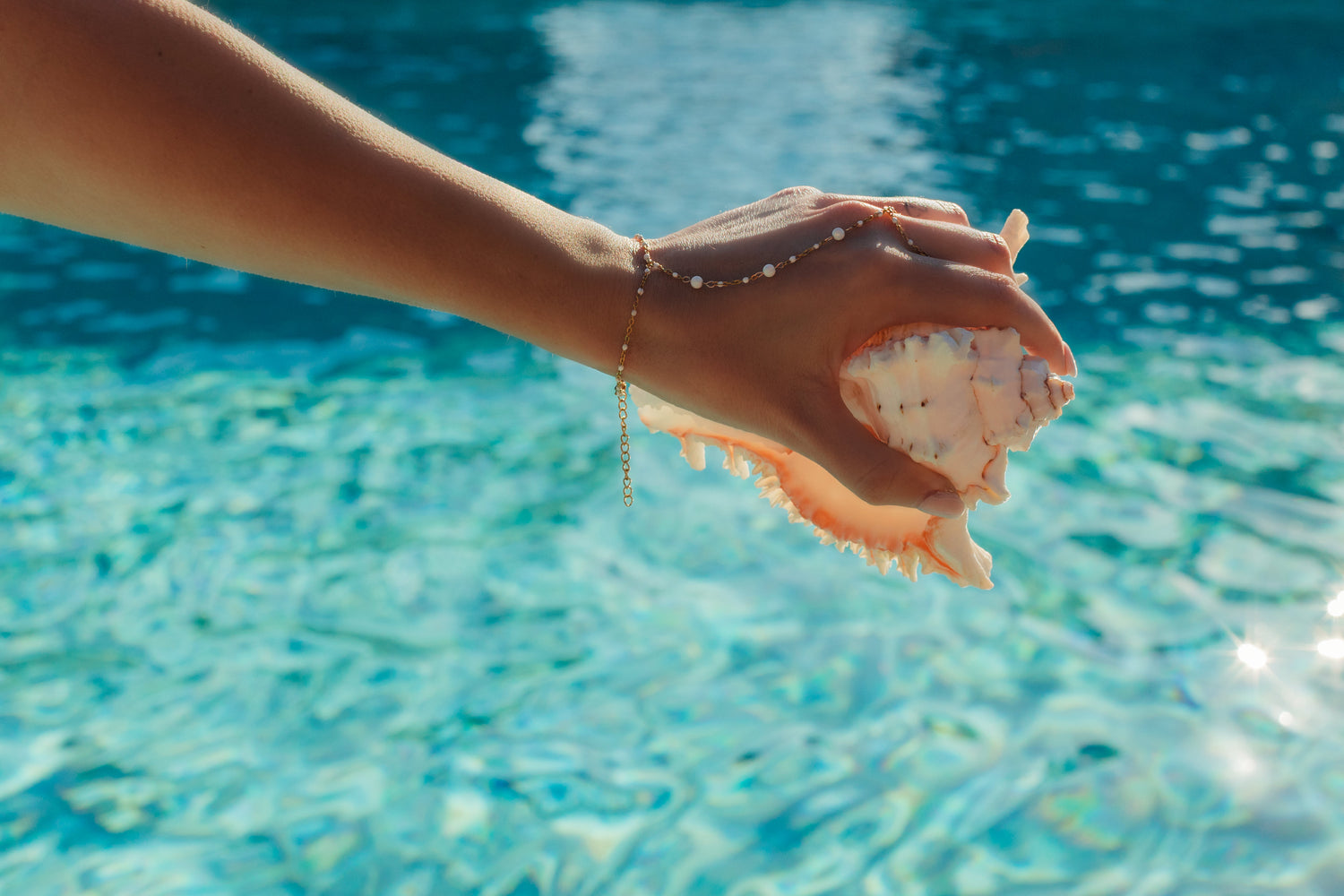 Hand holding a shell with a blurred pool background, wearing a watersafe coastal hand chain