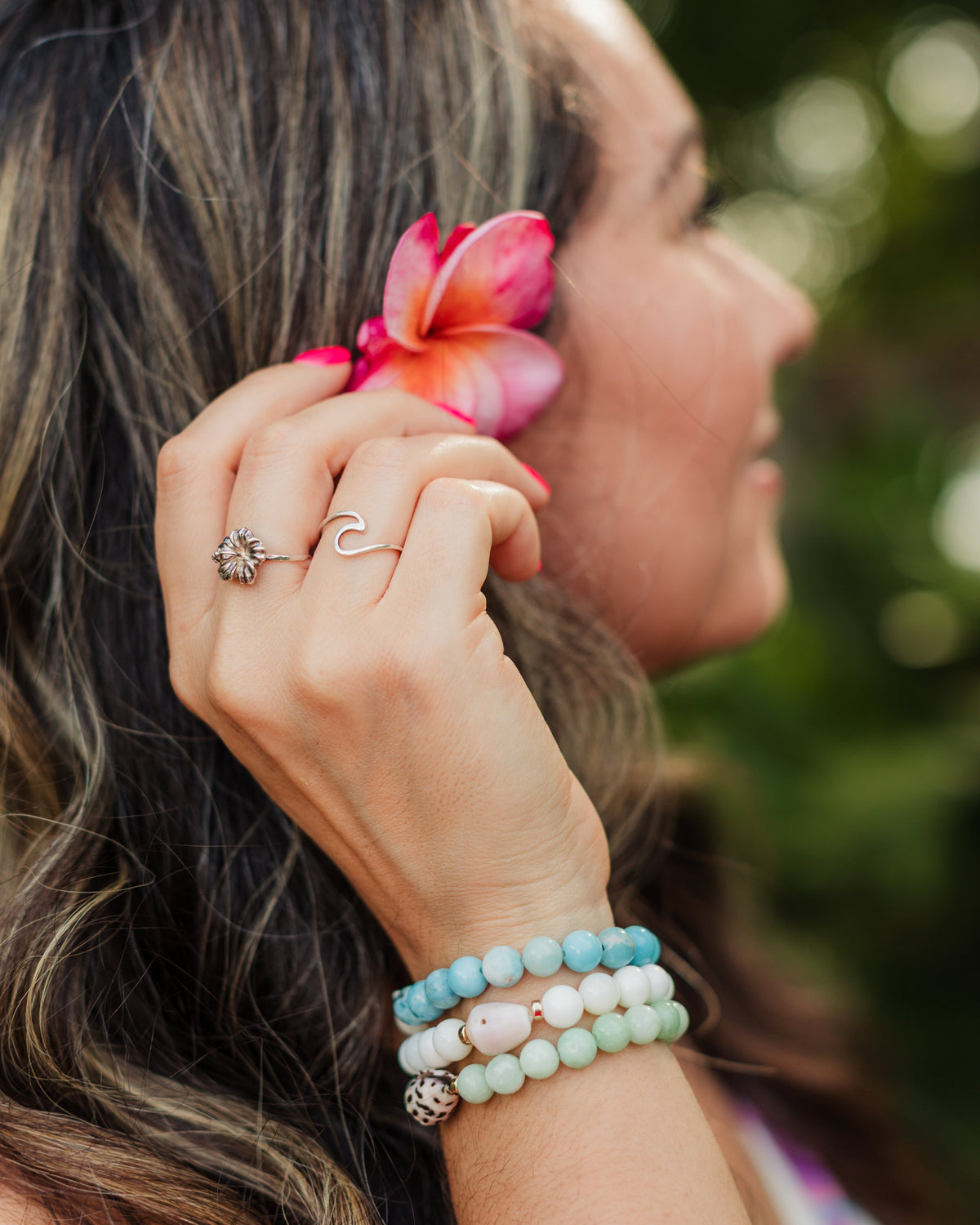 Woman with a pink flower in her hair and a stack of shell bracelets on her wrist, outdoors.