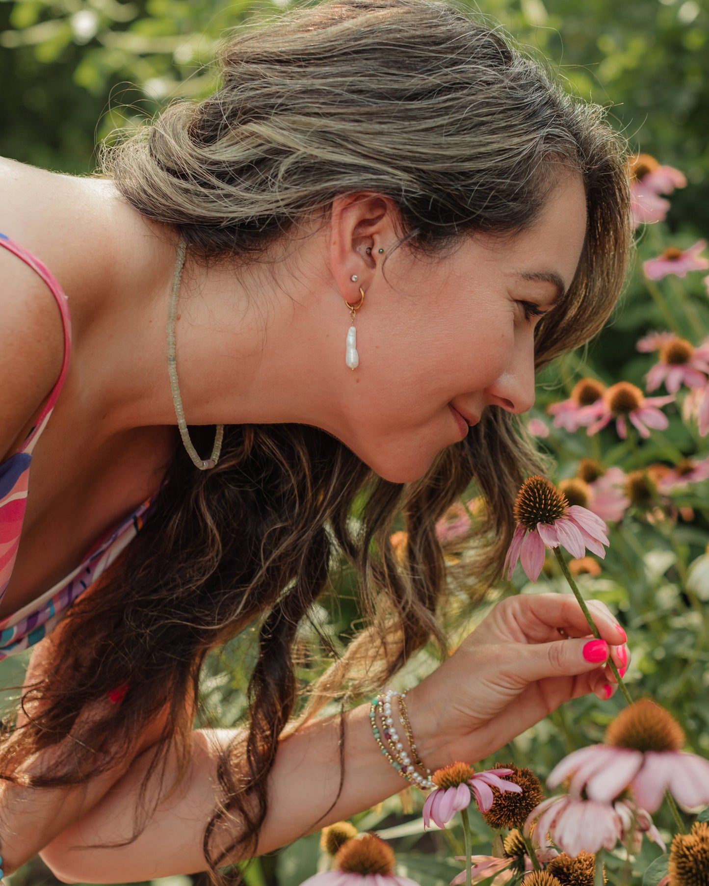Woman smelling a flower in a field of pink flowers wearing ocean pearl drop earrings. 