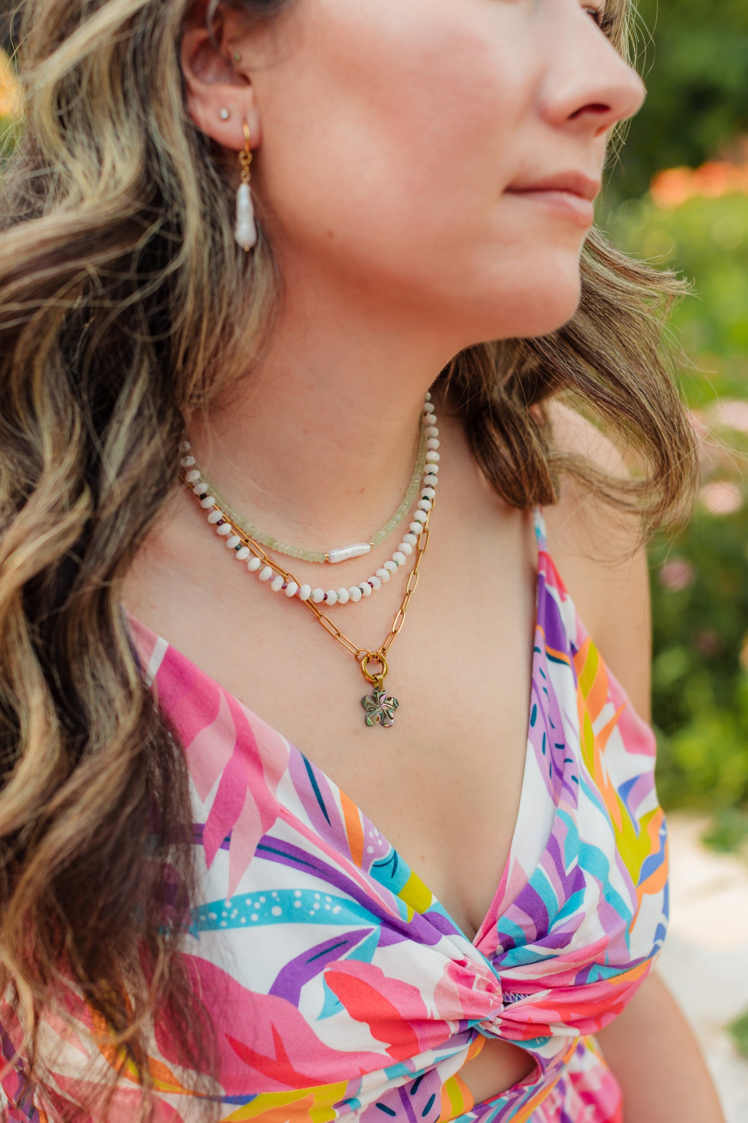 Woman wearing a colorful floral dress with layered necklaces and earrings outdoors.