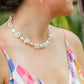 Woman in a colorful dress standing in a garden with orange flowers and greenery wearing Leilani flower pearl necklace.