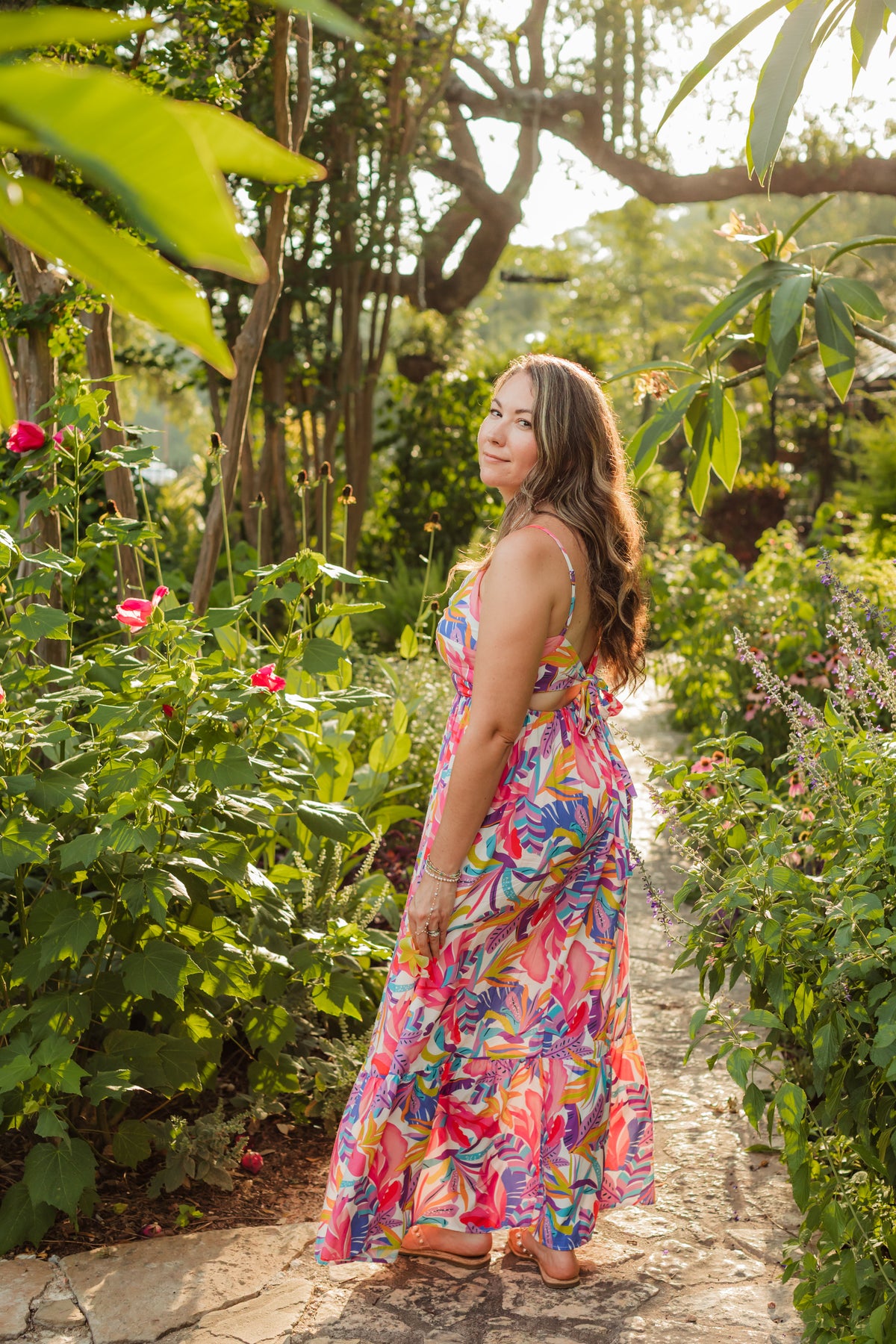 Woman in a colorful dress standing in a garden with flowers and trees.