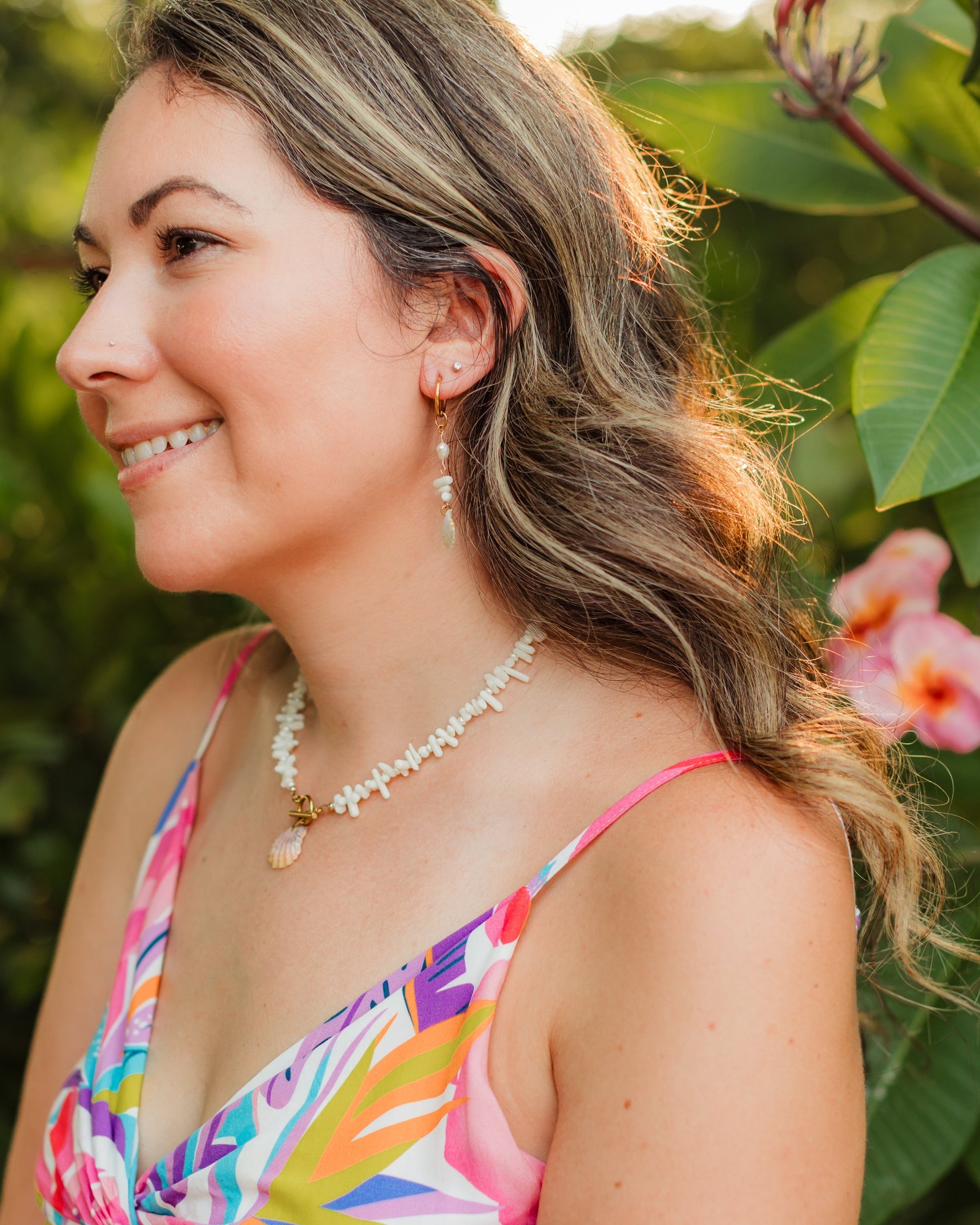 Woman in a colorful dress standing outdoors with greenery and flowers in the background wearing sunrise shell coral shell necklace and earrings.