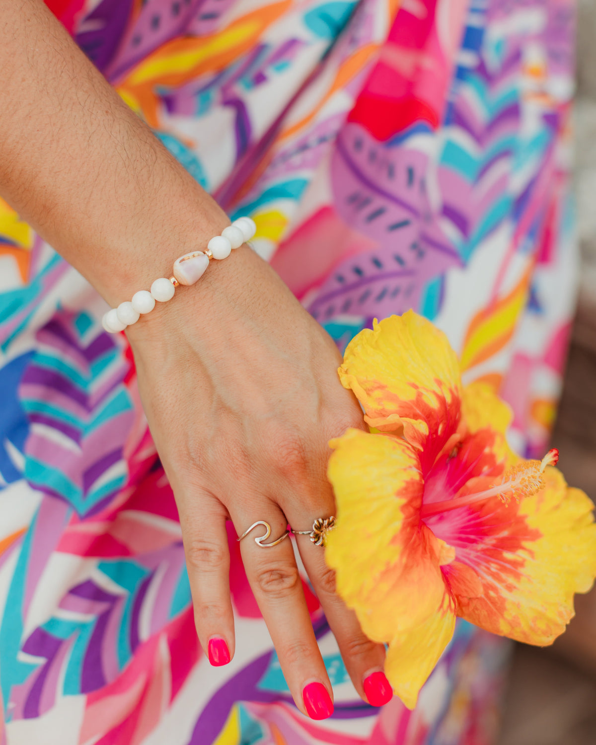 Person wearing a colorful dress holding a yellow flower wearing Lavender Bloom Bracelet with polished white shell beads, 14K gold-filled accents, and a rare natural lavender cone shell
