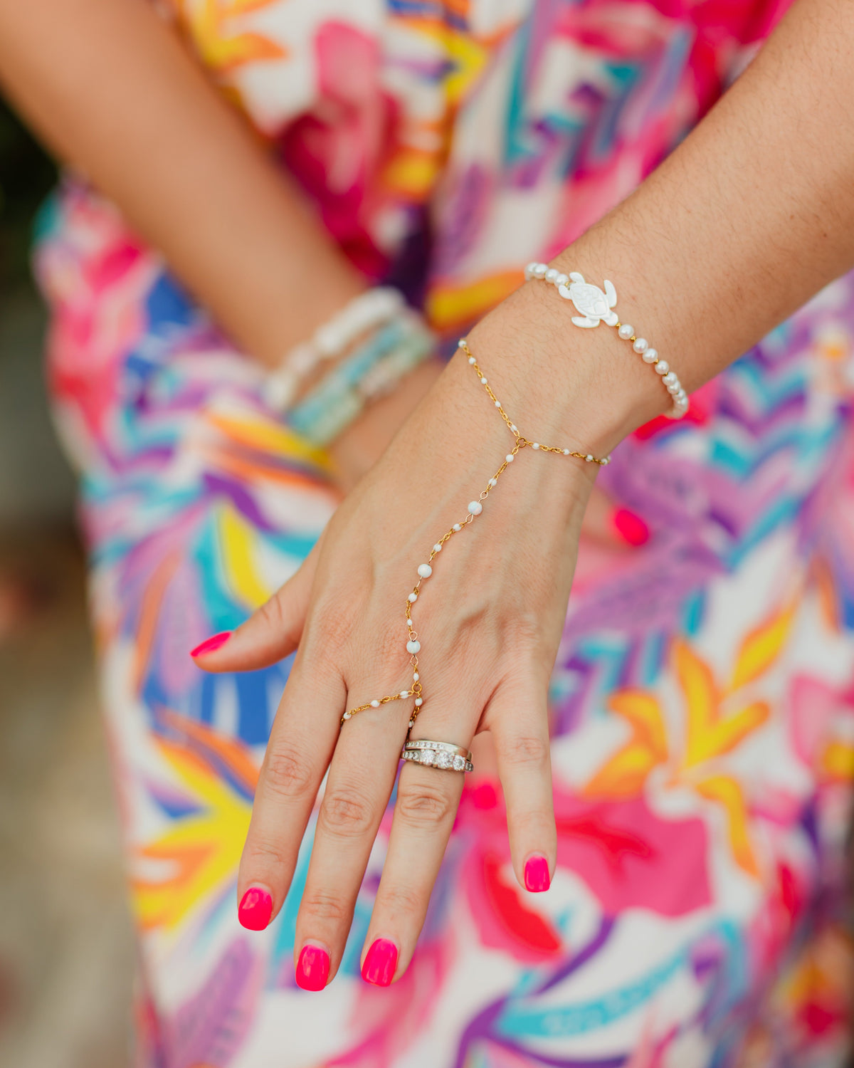 Close-up of a person's hand wearing multiple bracelets with a colorful dress in the background honu bracelet and hand chain