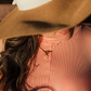 Person wearing a wide-brimmed hat and pink top with a necklace, blurred background, wearing island blush plumeria necklace