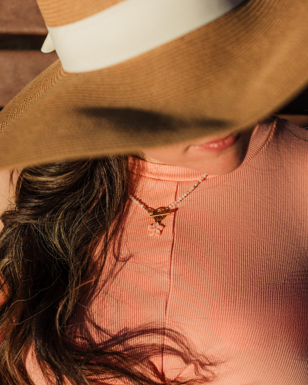 Person wearing a wide-brimmed hat and pink top with a necklace, blurred background, wearing island blush plumeria necklace