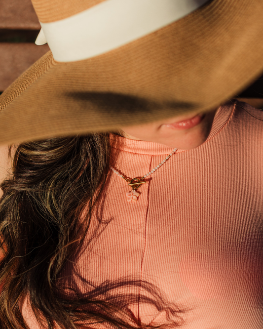 Person wearing a wide-brimmed hat and pink top with a necklace, blurred background, wearing island blush plumeria necklace