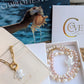 A jewelry set from Charlotte's Cove, featuring a gold chain necklace with a white pearl flower charm and a multi-strand pearl bracelet with a silver angel charm, displayed in their white gift boxes with the brand's packaging in the background.