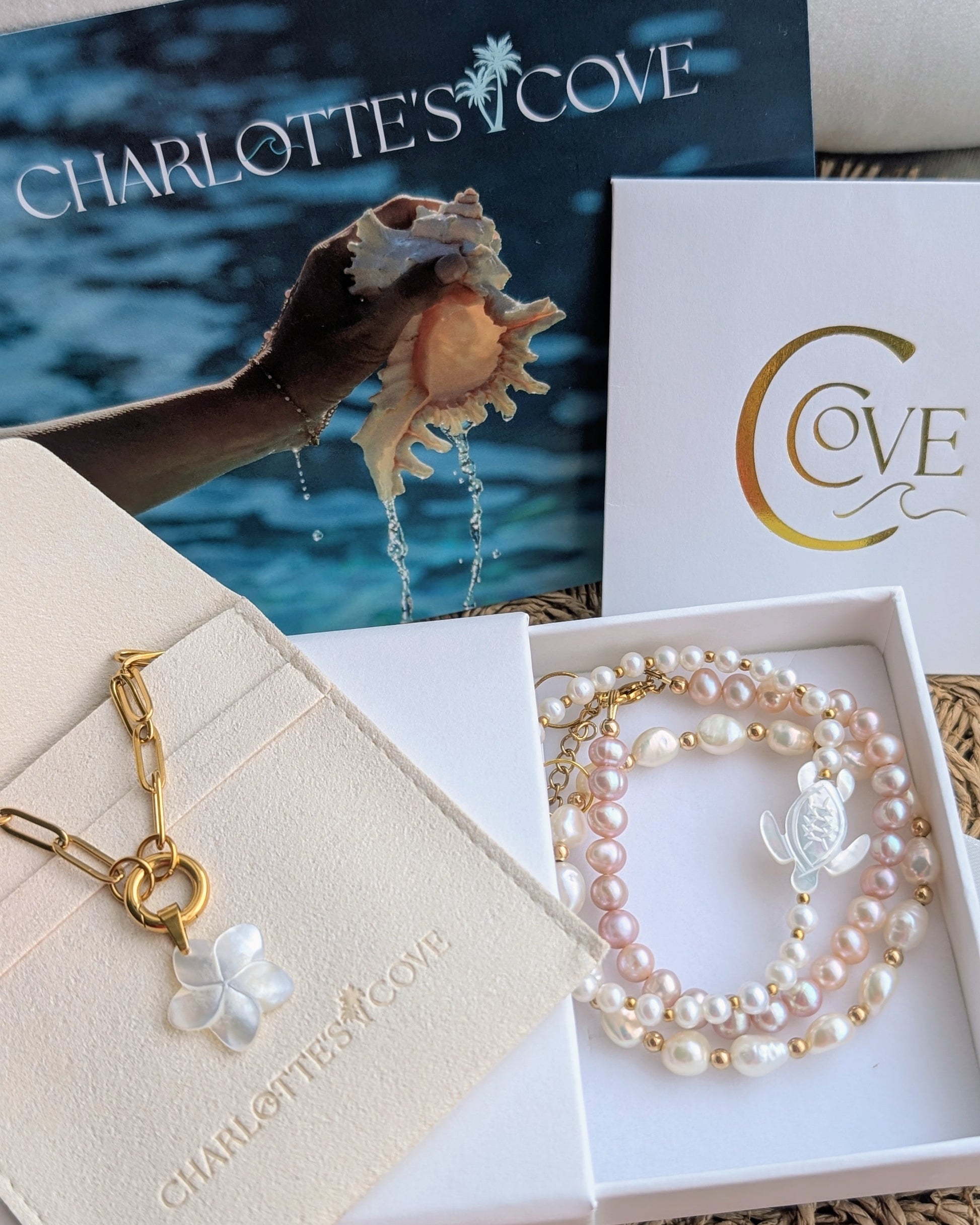 A jewelry set from Charlotte's Cove, featuring a gold chain necklace with a white pearl flower charm and a multi-strand pearl bracelet with a silver angel charm, displayed in their white gift boxes with the brand's packaging in the background.
