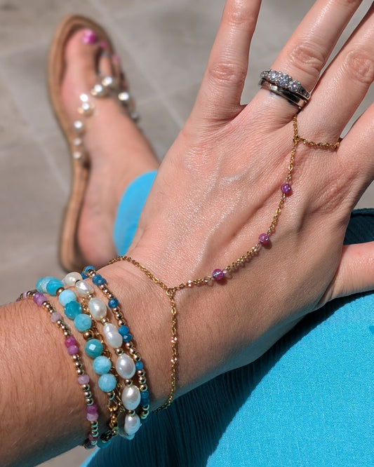 A close-up of a person's hand and wrist wearing a delicate gold chain hand bracelet with small purple and blue beads, along with pearl and beaded wristbands and a silver ring. The person is wearing bright pink nail polish and tan flat sandals, resting their hand on a blue surface outdoors.