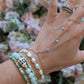 A close-up of a person's hand and wrist wearing multiple delicate jewelry pieces: a gold chain hand chain with small turquoise beads, a stack of beaded bracelets in mint green, white, and a cowrie shell, and stacked rings on the index finger, set against a soft-focus background of pink and white flowers.