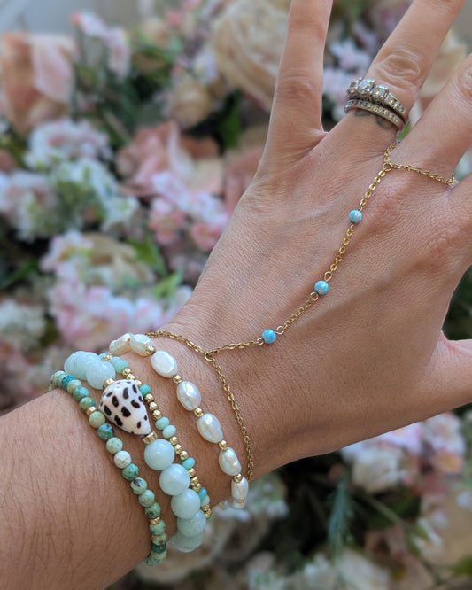 A close-up of a person's hand and wrist wearing multiple delicate jewelry pieces: a gold chain hand chain with small turquoise beads, a stack of beaded bracelets in mint green, white, and a cowrie shell, and stacked rings on the index finger, set against a soft-focus background of pink and white flowers.