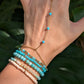 A gold chain hand chain bracelet with small turquoise blue beads worn on a person's wrist and hand, paired with multiple beaded bracelets, set against a background of large green leaves.