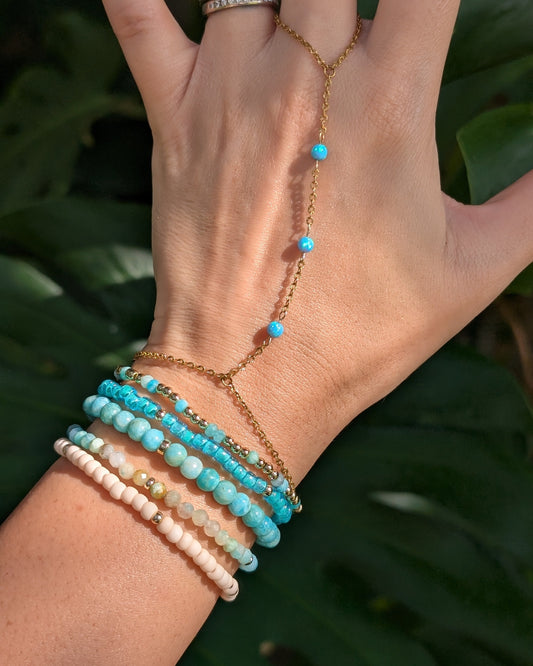 A gold chain hand chain bracelet with small turquoise blue beads worn on a person's wrist and hand, paired with multiple beaded bracelets, set against a background of large green leaves.