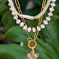 A layered gold-tone necklace featuring white pearl-like beads, a baroque pearl, and a dangling four-leaf clover charm, displayed against a blurred background of green leaves.