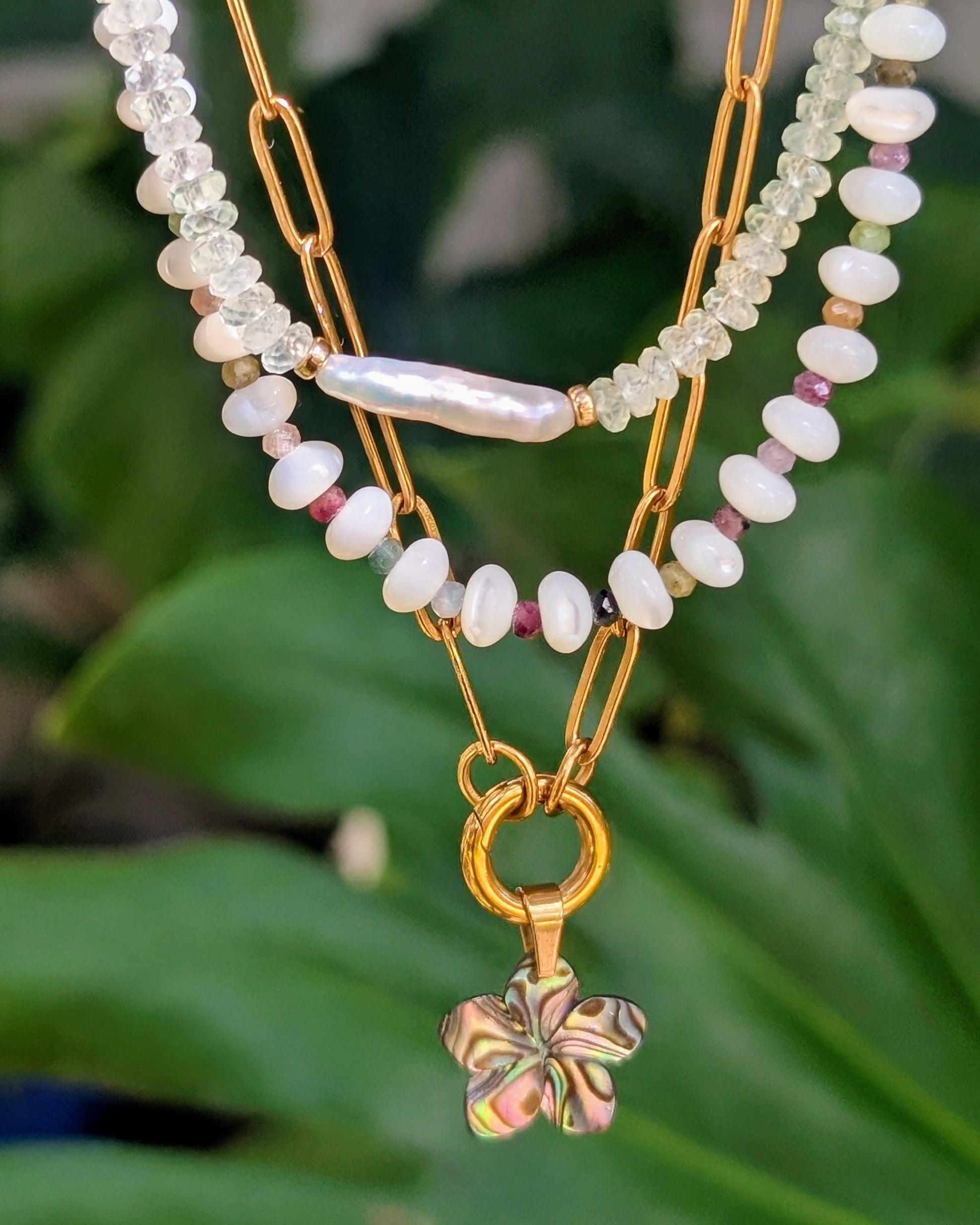 A layered gold-tone necklace featuring white pearl-like beads, a baroque pearl, and a dangling four-leaf clover charm, displayed against a blurred background of green leaves.