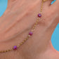 A gold chain anklet with three faceted magenta-purple beads, shown worn on a person's ankle with a blue background.