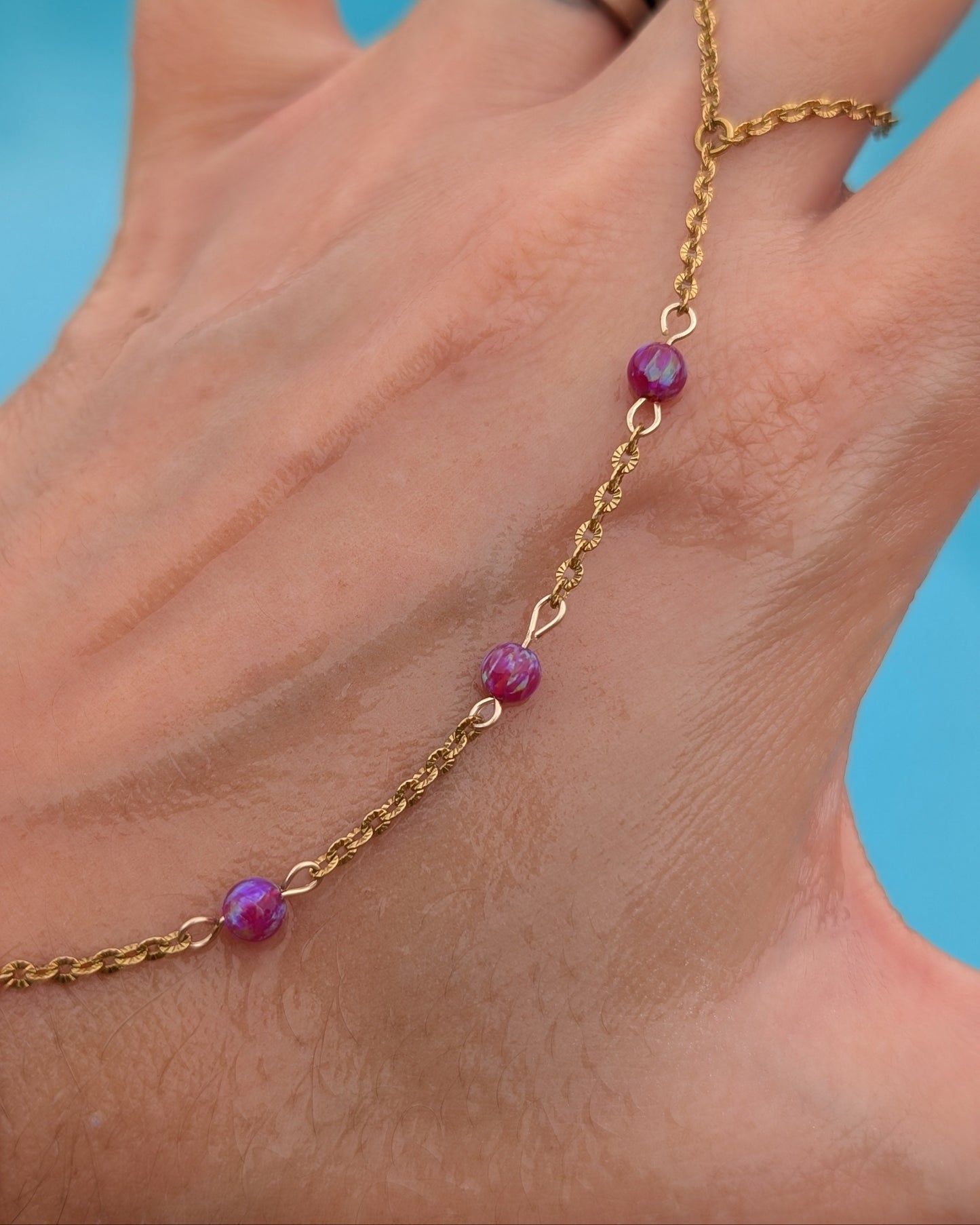 A gold chain anklet with three faceted magenta-purple beads, shown worn on a person's ankle with a blue background.