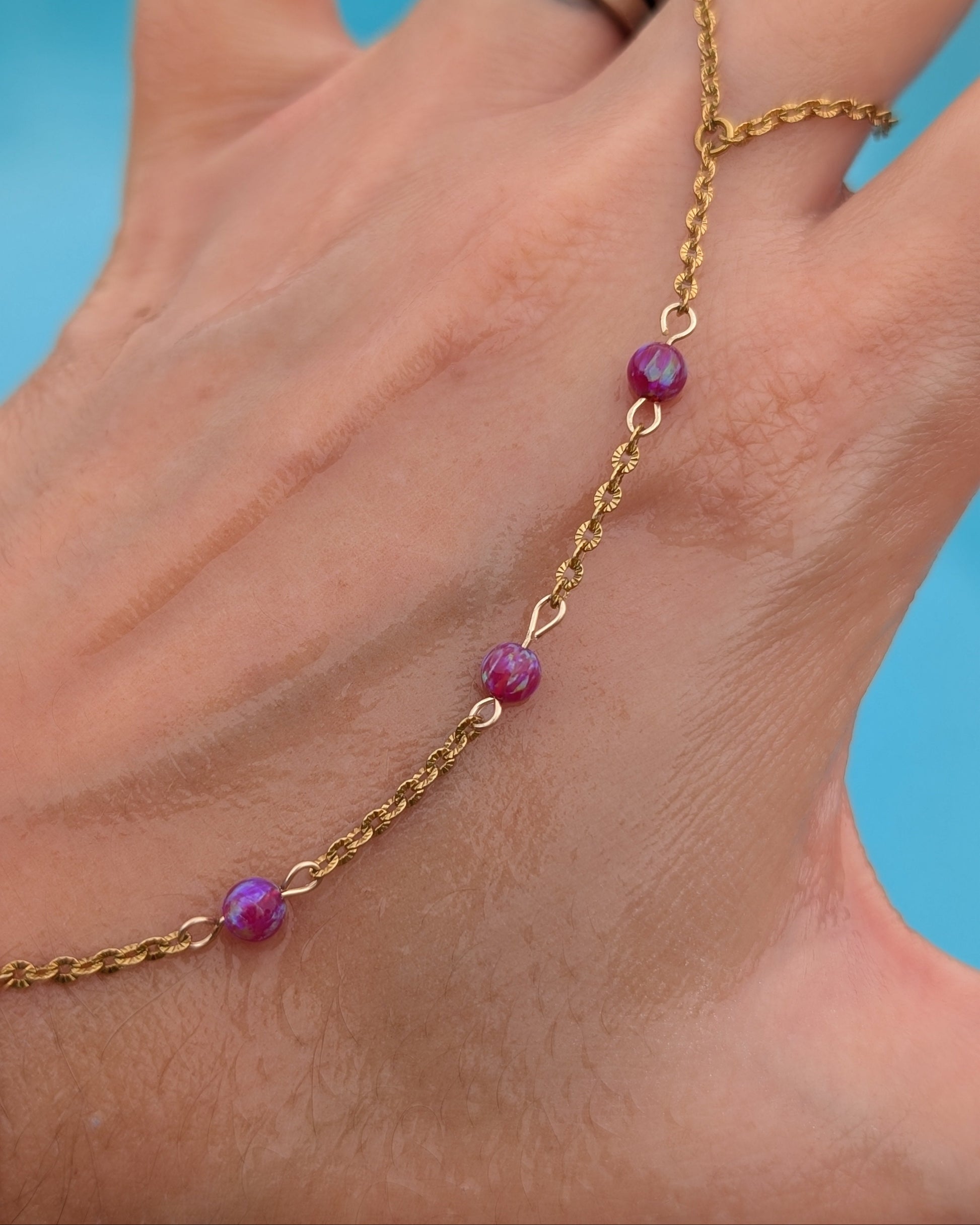 A gold chain anklet with three faceted magenta-purple beads, shown worn on a person's ankle with a blue background.