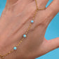 A gold chain hand bracelet with small light blue opal-like beads, shown worn on a person's hand against a bright blue background. The person is also wearing two silver rings on their fingers.
