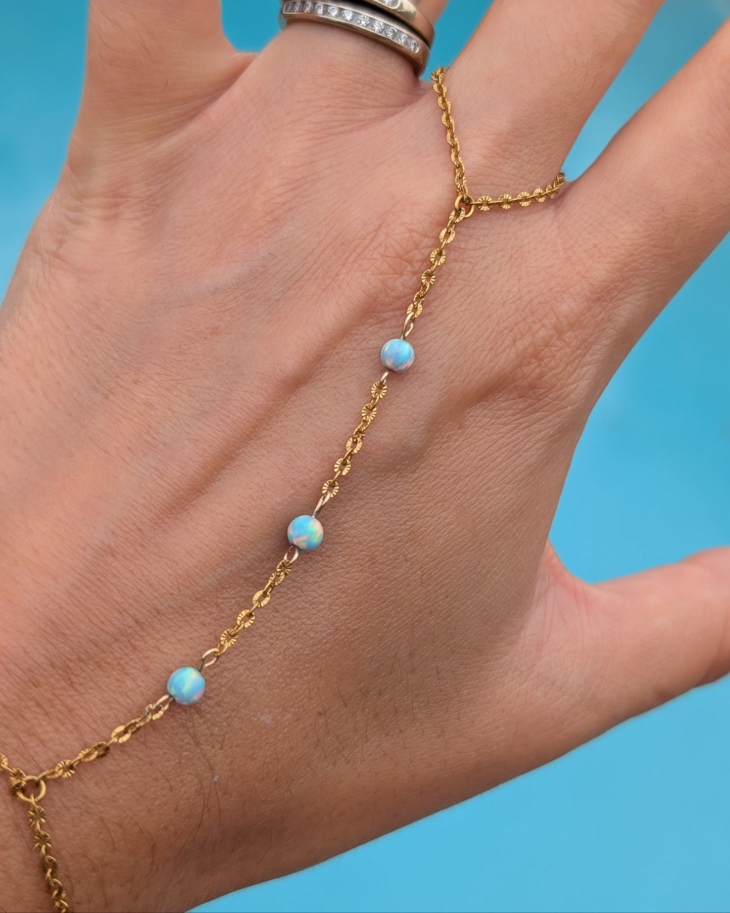 A gold chain hand bracelet with small light blue opal-like beads, shown worn on a person's hand against a bright blue background. The person is also wearing two silver rings on their fingers.