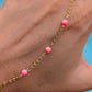 A close-up of a hand wearing a delicate gold chain anklet decorated with four small pink marbled beads, set against a bright blue background.