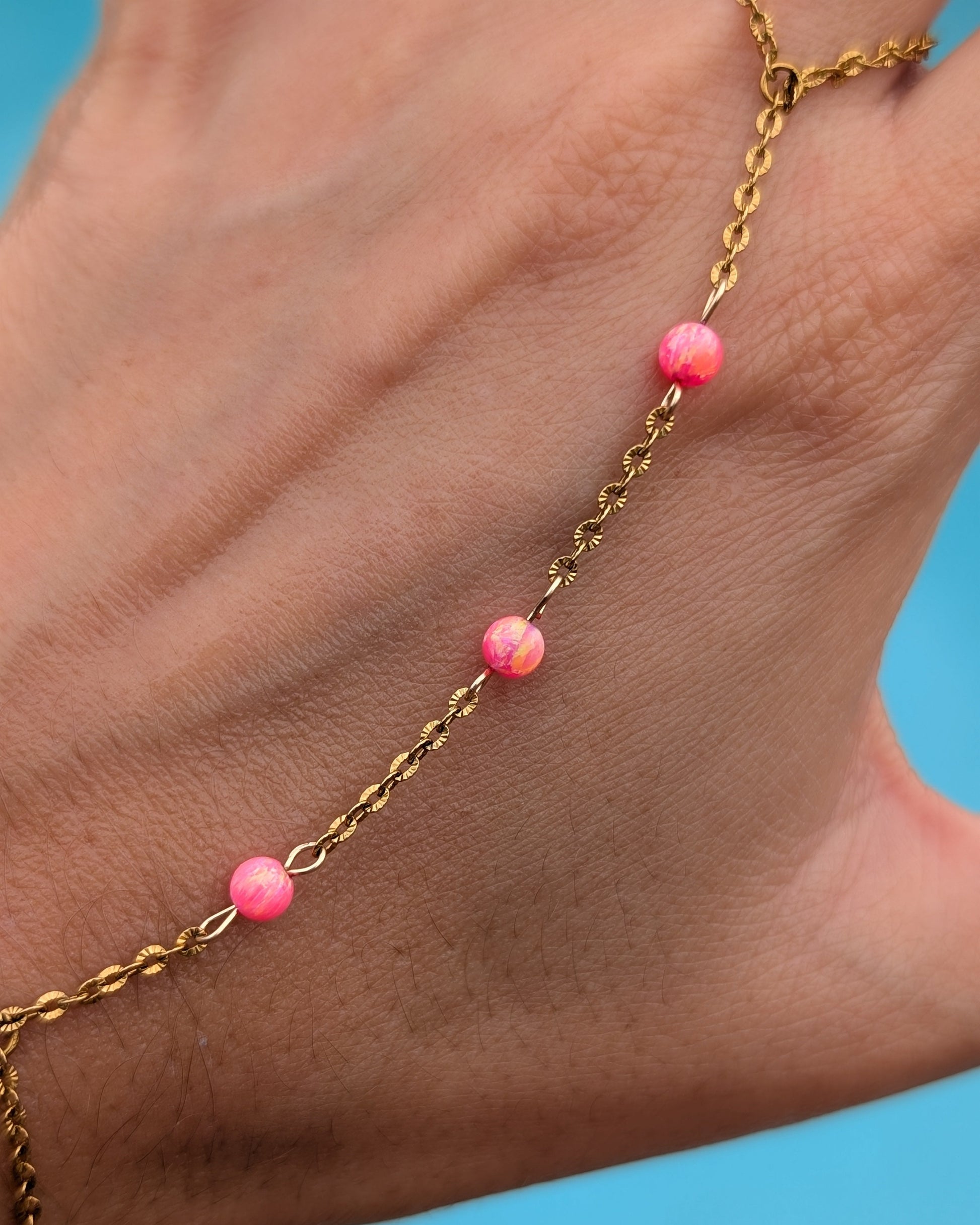 A close-up of a hand wearing a delicate gold chain anklet decorated with four small pink marbled beads, set against a bright blue background.