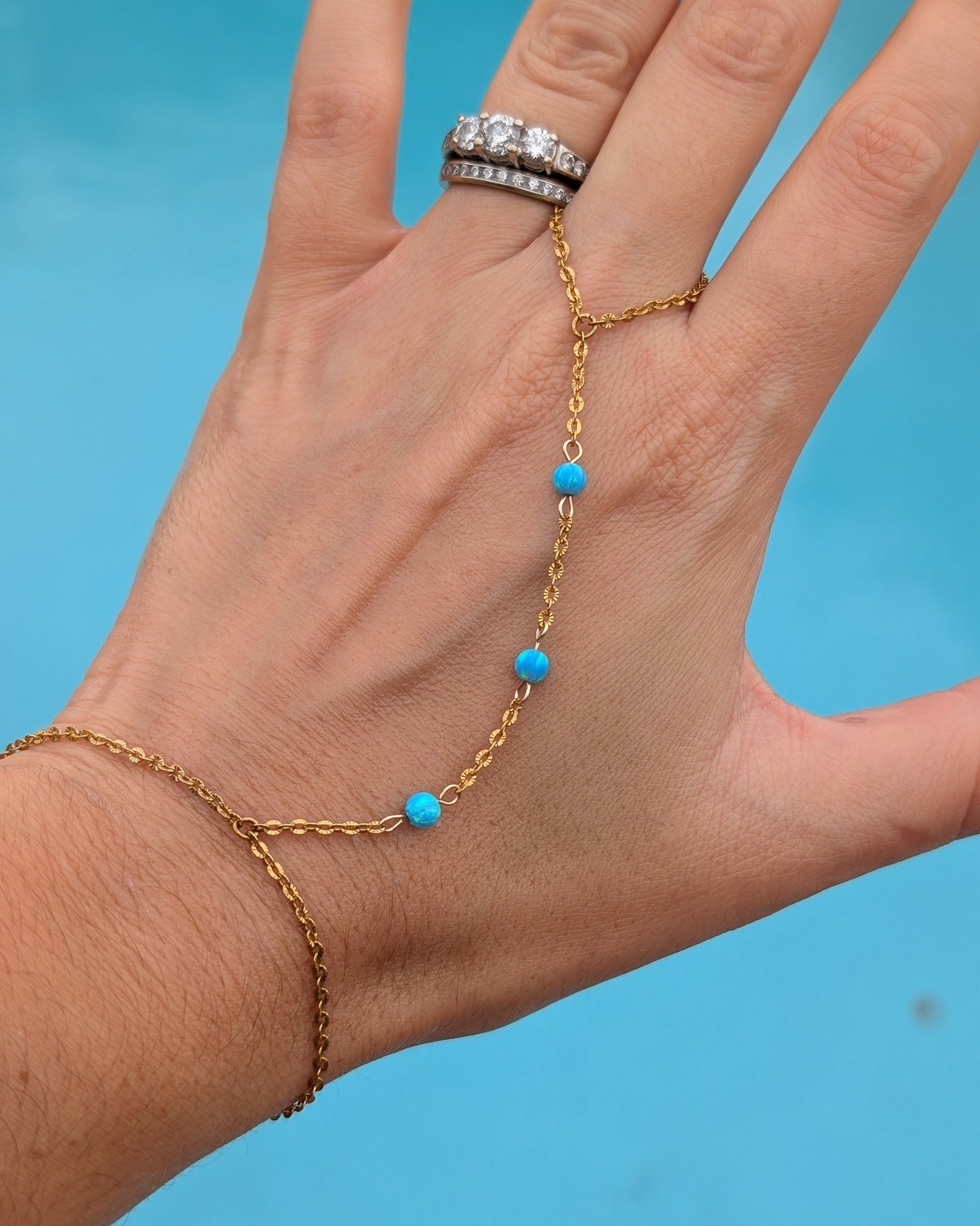 A gold chain hand and wrist bracelet with small turquoise blue beads, shown worn on a person's hand against a bright blue background.