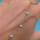 A gold chain anklet with four small light blue opal-like beads worn on a person's ankle, shown against a bright blue background.