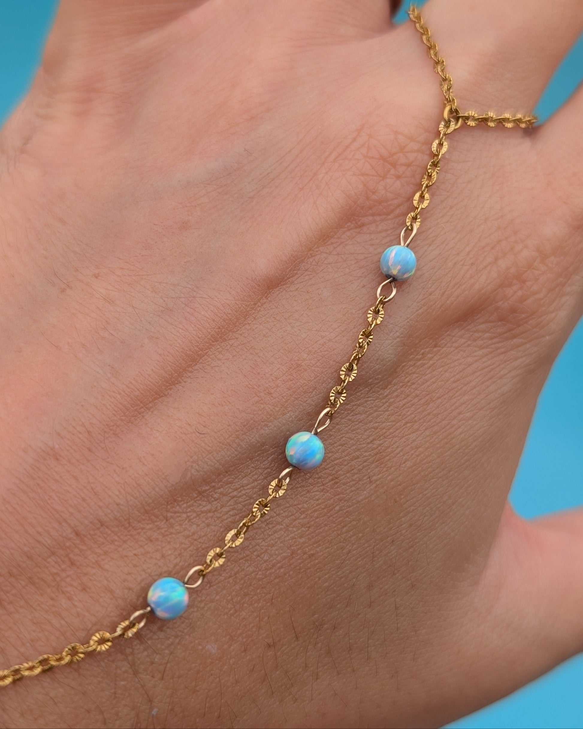 A gold chain anklet with four small light blue opal-like beads worn on a person's ankle, shown against a bright blue background.