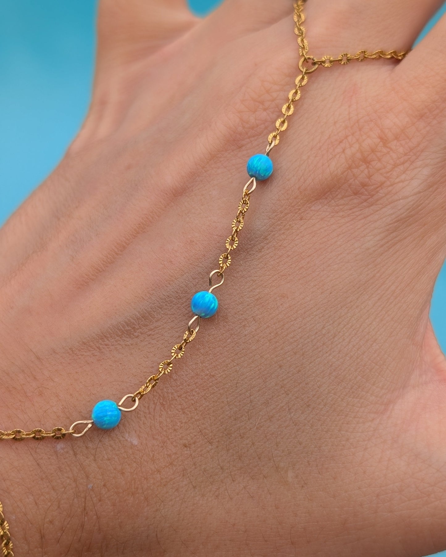 A close-up of a hand wearing a delicate gold chain anklet with three small bright turquoise blue beads spaced along the chain.