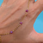 A gold chain anklet with three small purple beads worn on a person's ankle, shown against a bright blue background.