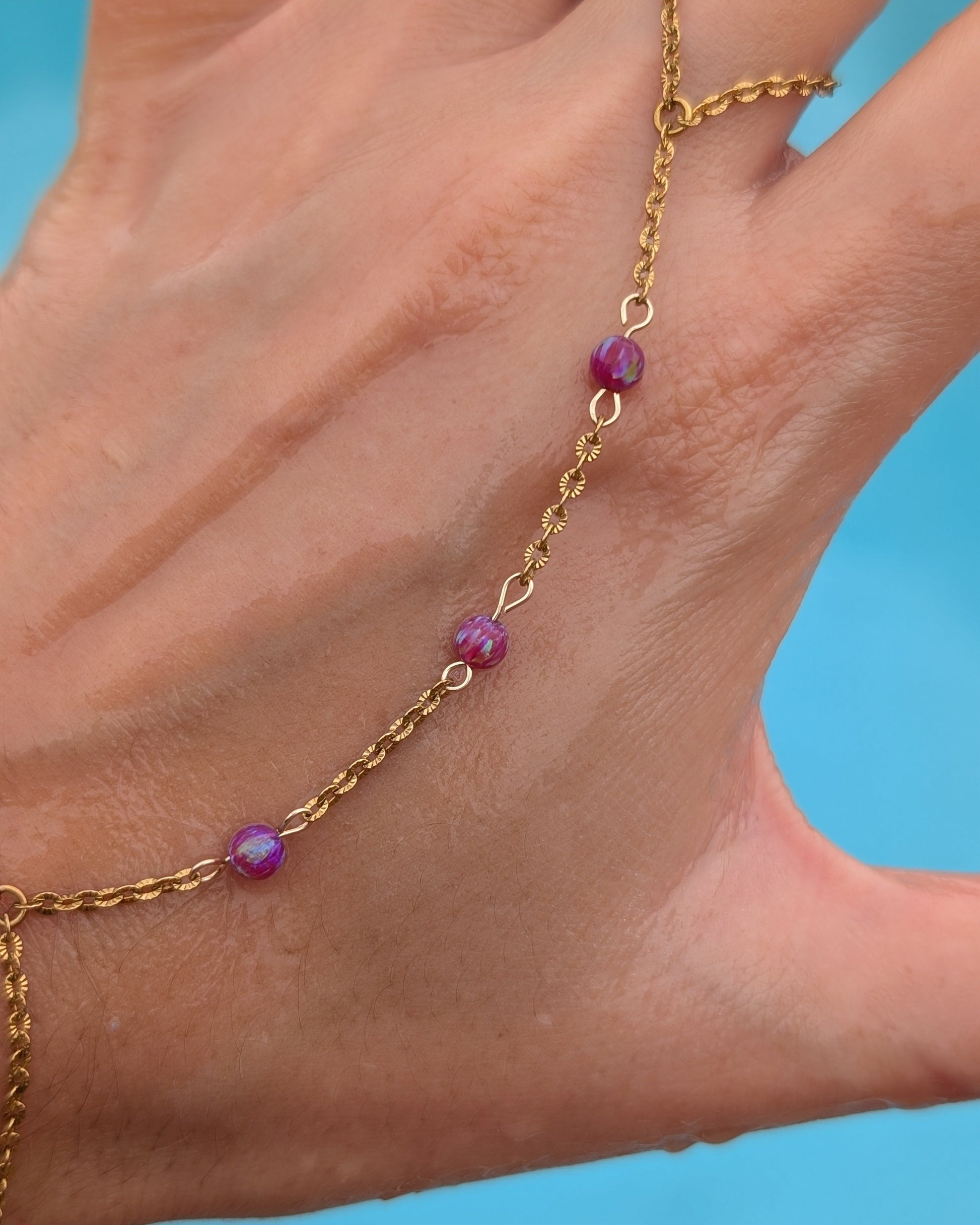 A gold chain anklet with three small purple beads worn on a person's ankle, shown against a bright blue background.