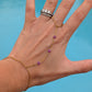 A gold chain hand bracelet with small purple bead accents, shown worn on a person's hand with a silver ring on the finger, against a bright blue background.