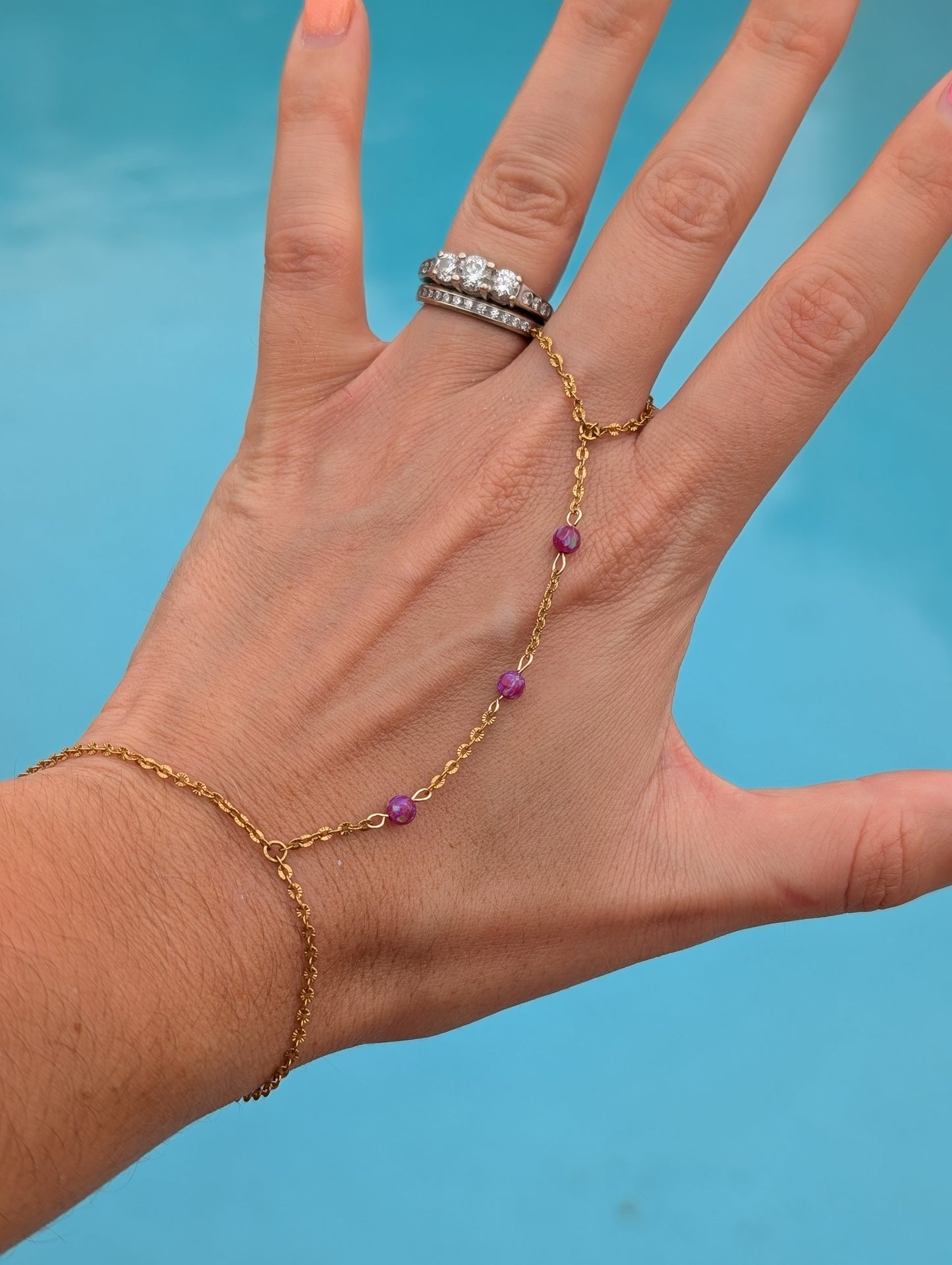 A gold chain hand bracelet with small purple bead accents, shown worn on a person's hand with a silver ring on the finger, against a bright blue background.