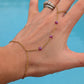 A gold chain hand bracelet with small purple gemstone beads worn on a person's wrist, shown against a bright blue background. The person's hand is also wearing a silver ring.