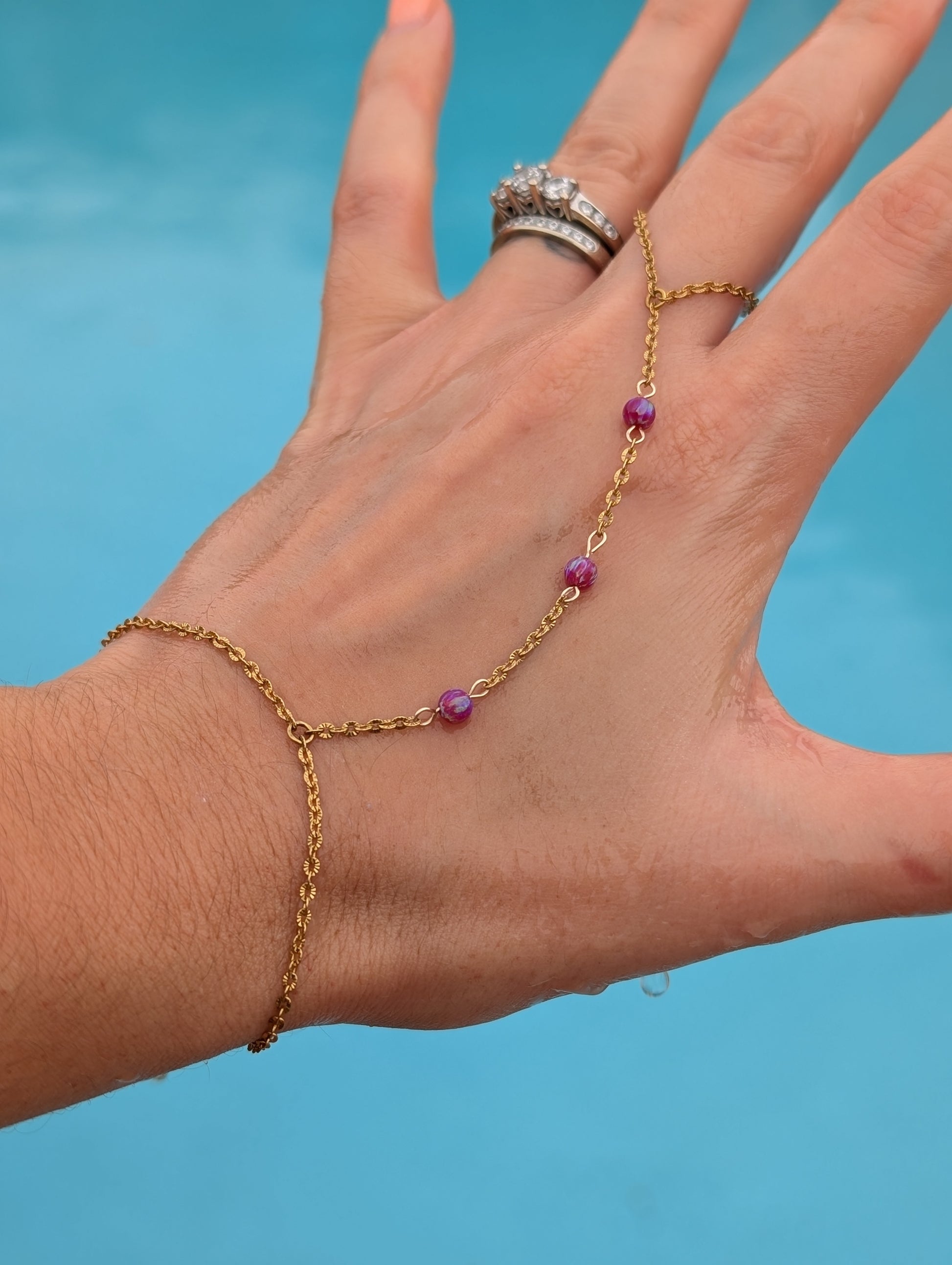 A gold chain hand bracelet with small purple gemstone beads worn on a person's wrist, shown against a bright blue background. The person's hand is also wearing a silver ring.