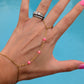 A gold chain hand bracelet with small pink beads draped across a hand, shown against a bright blue background. The hand also has a silver ring with clear stones.