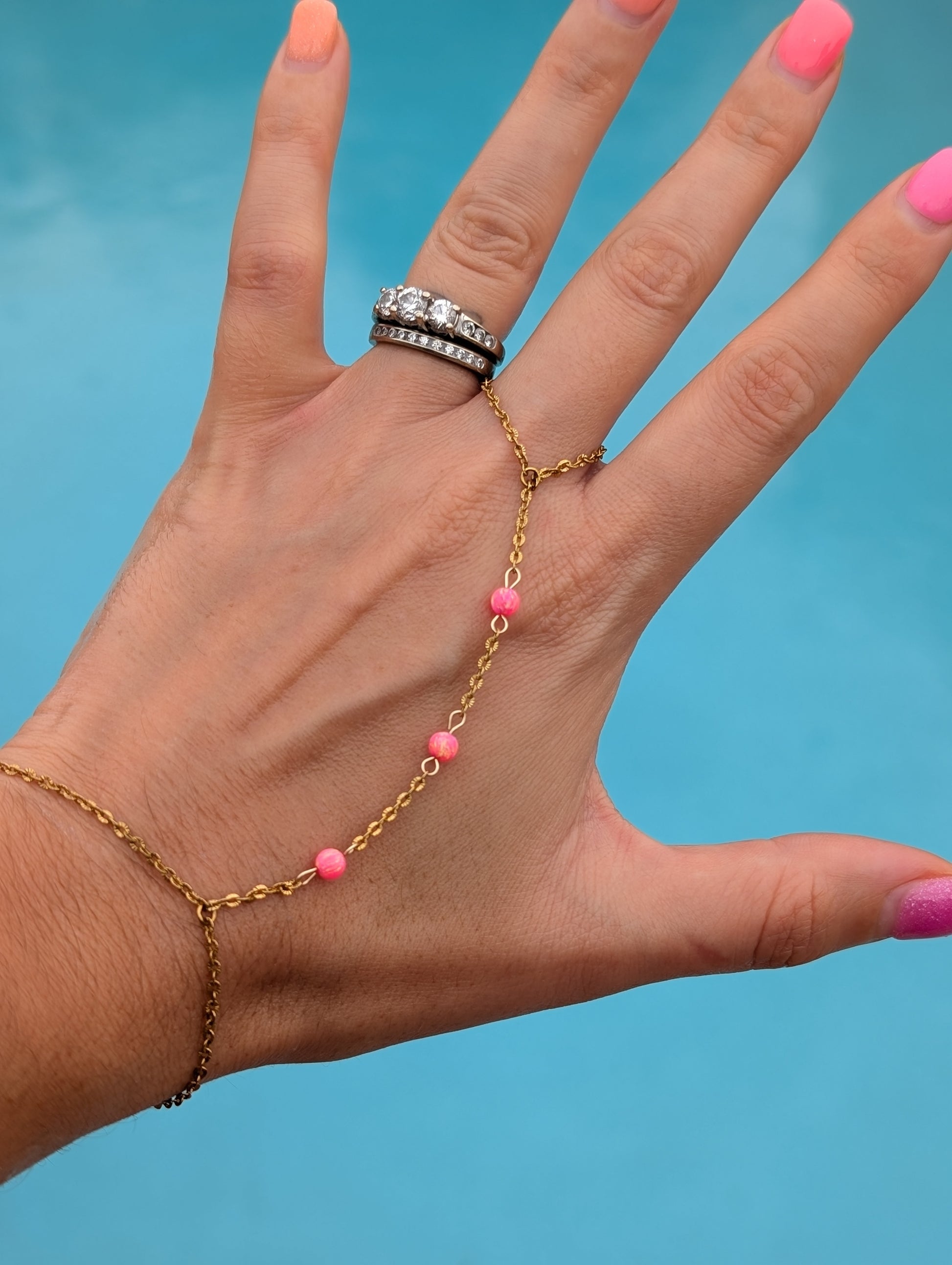 A gold chain hand bracelet with small pink beads draped across a hand, shown against a bright blue background. The hand also has a silver ring with clear stones.