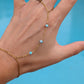 A gold chain anklet with three small light blue beads worn on a person's ankle and wrist, shown on a hand against a bright blue background.