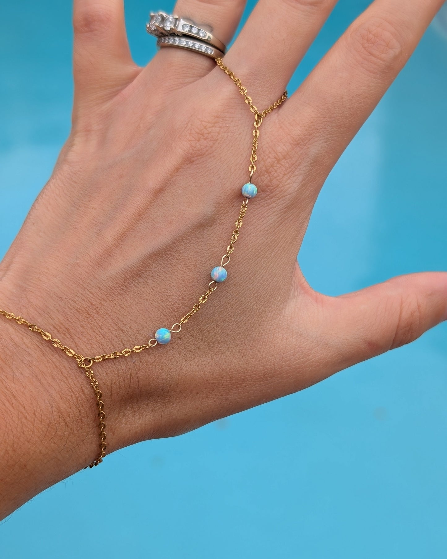 A gold chain anklet with three small light blue beads worn on a person's ankle and wrist, shown on a hand against a bright blue background.