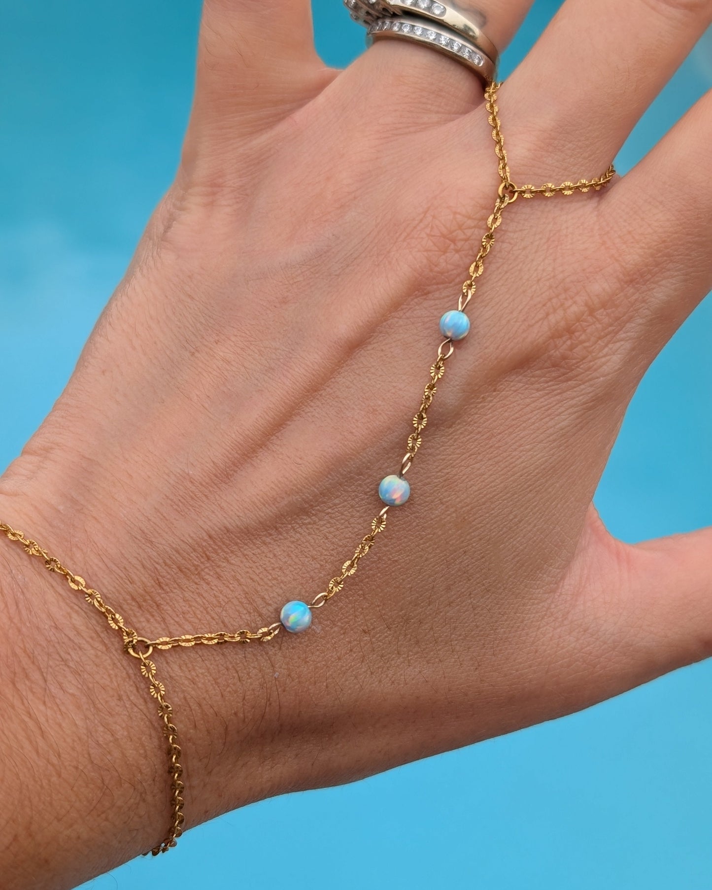 A gold chain hand bracelet with four small light blue beads, shown worn on a hand against a bright blue background.