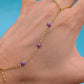 A gold chain hand bracelet with three small purple and pink iridescent beads, shown worn on a hand against a bright blue background.